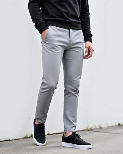 Hellgraue Chino im Freien vor einer weißen Wand, kombiniert mit schwarzem Pullover und schwarzen Sneakern mit weißer Sohle. 2