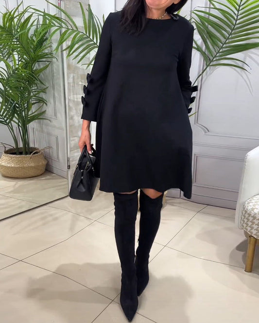 Frau in schwarzem Kleid und Stiefeln, hält schwarze Handtasche, steht in modernem Raum.