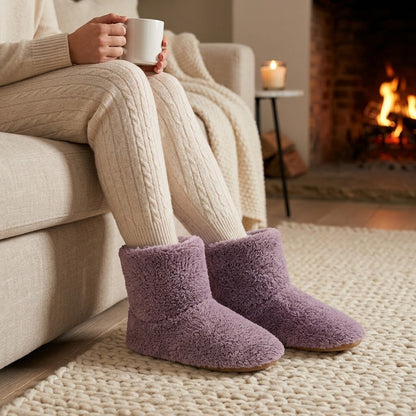 Person in cremefarbener Strickhose hält am Sofa eine Tasse und trägt flauschige Plüsch-Stiefelhausschuhe in Flieder vor dem Kamin. 2