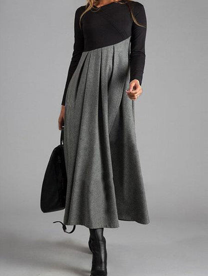 Frau in langem, grauem Maxikleid mit schwarzen Ärmeln und Stiefeln, hält schwarze Tasche.