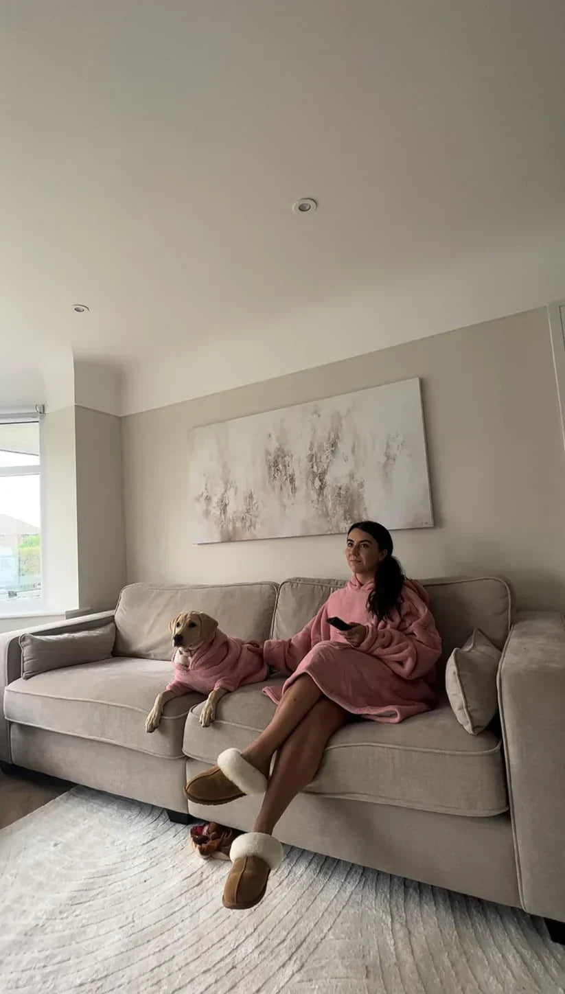 Frau und Hund in rosa Decken auf grauem Sofa, gemütliches Wohnzimmer, entspannte Atmosphäre.