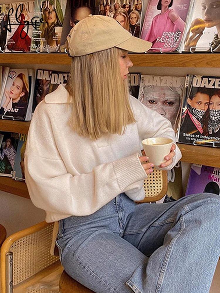 Frau in weißem Pullover und Jeans, beige Kappe, hält Kaffeetasse, sitzt vor Zeitschriftenregal.