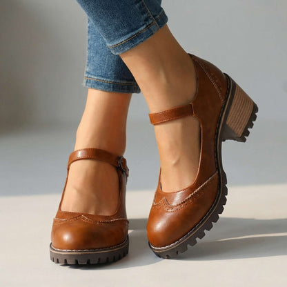 Braune Mary Janes mit Lochmuster und Riemen am Knöchel, am Fuß mit Jeans vor hellem Hintergrund, Frontansicht. 2