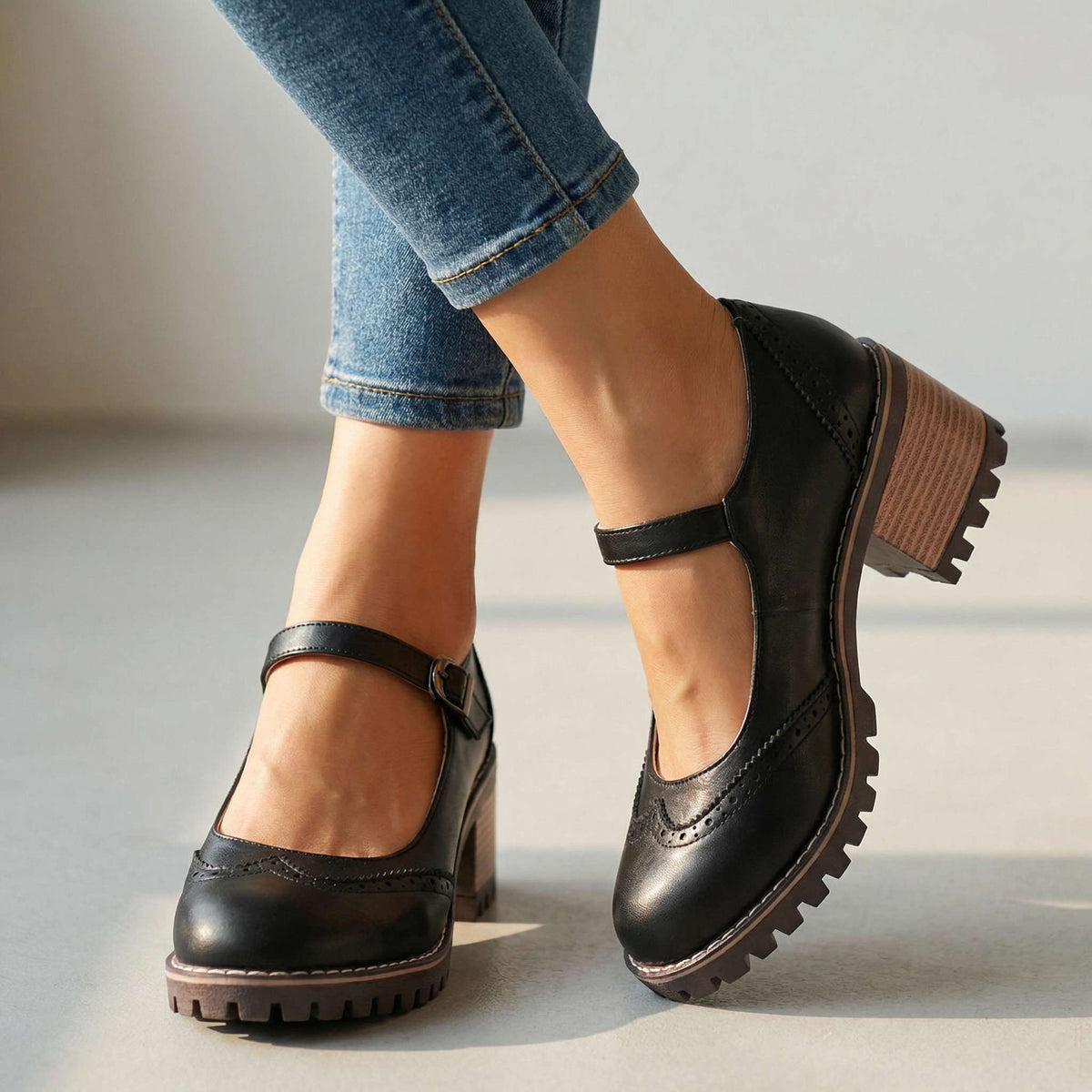 Schwarze Mary Janes mit Lochmuster, Knöchelriemen und Blockabsatz, getragen mit Jeans vor hellem Studiohintergrund, von vorne gesehen. 1