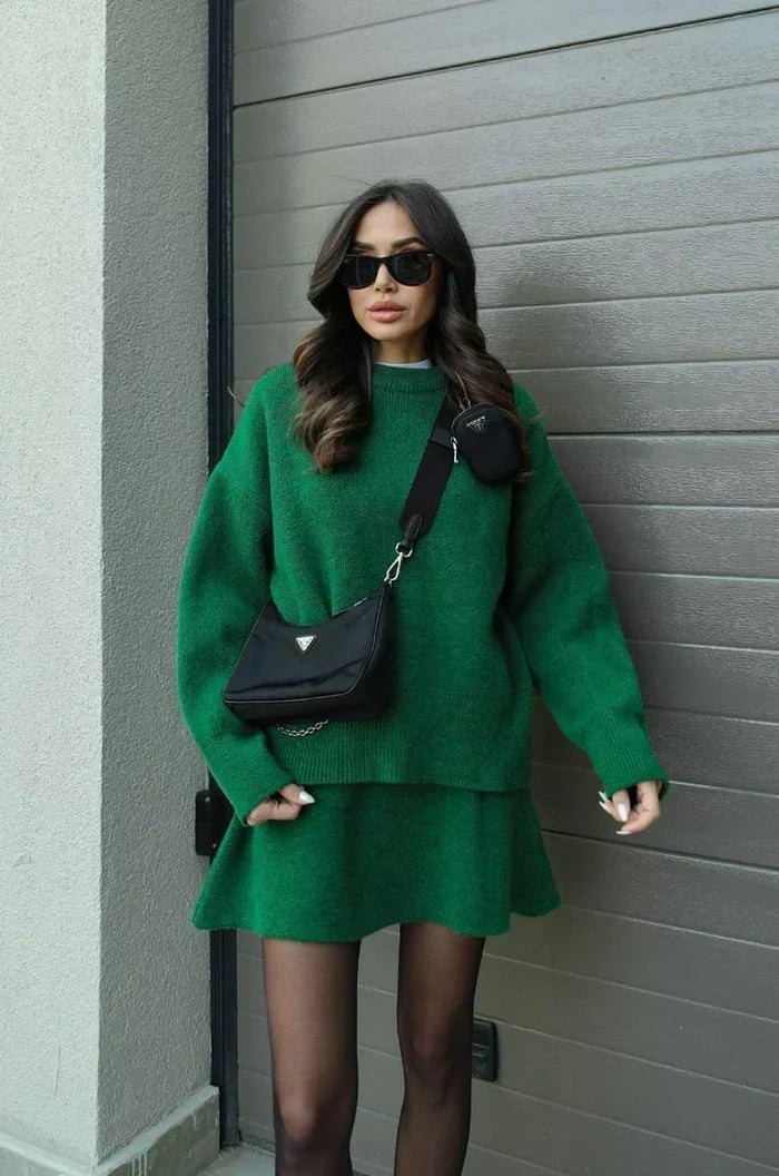 Model in grünem Strick-Set aus Pullover und ausgestelltem Minirock, mit schwarzer Umhängetasche vor einem Garagentor. 8