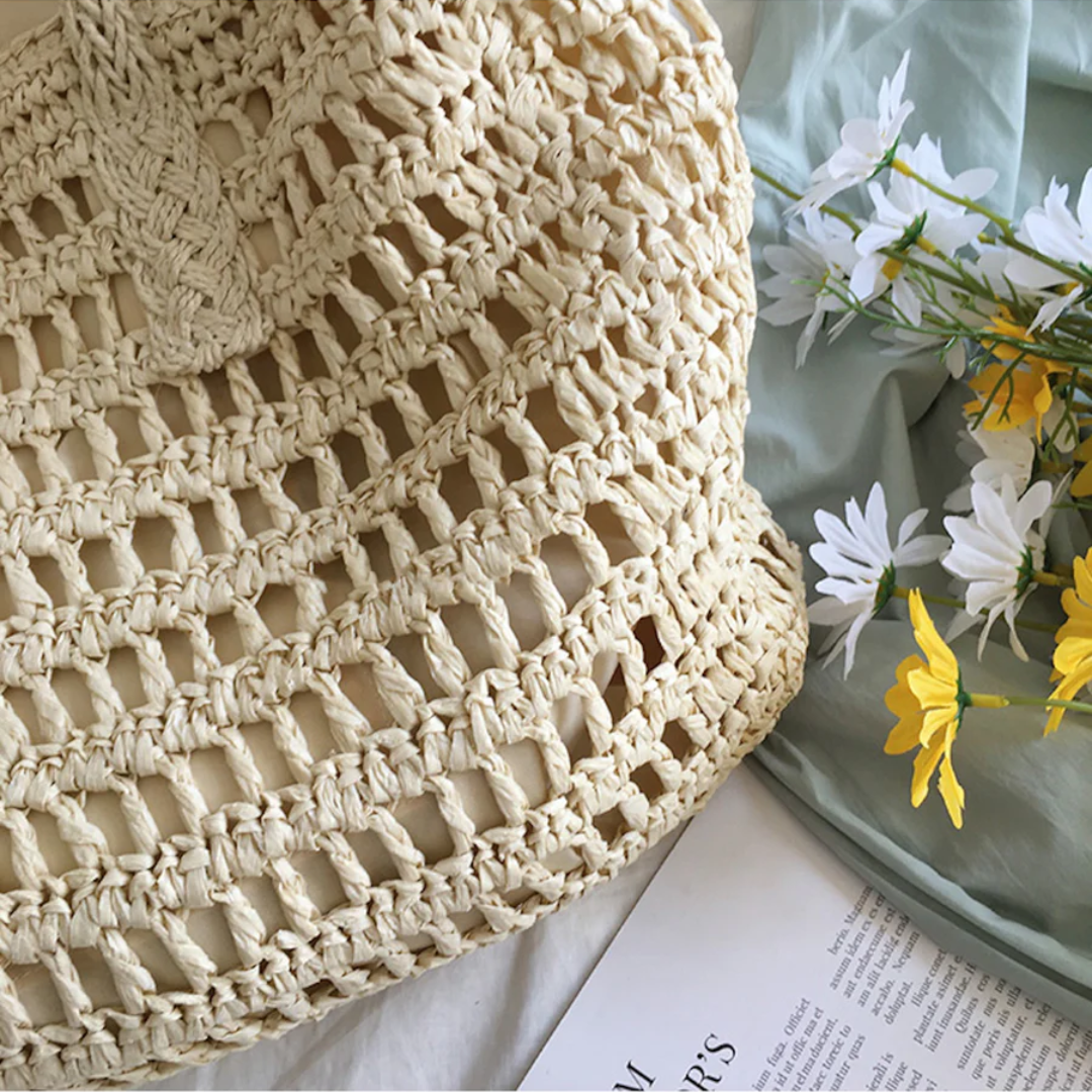 Beige gehäkelte Tasche aus Stroh neben weißen und gelben Blumen auf grünem Stoff.