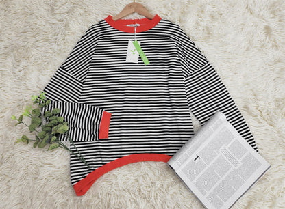 Schwarz-weiß gestreifter Pullover mit roten Bündchen auf Teppich, neben Zeitung und Pflanze.