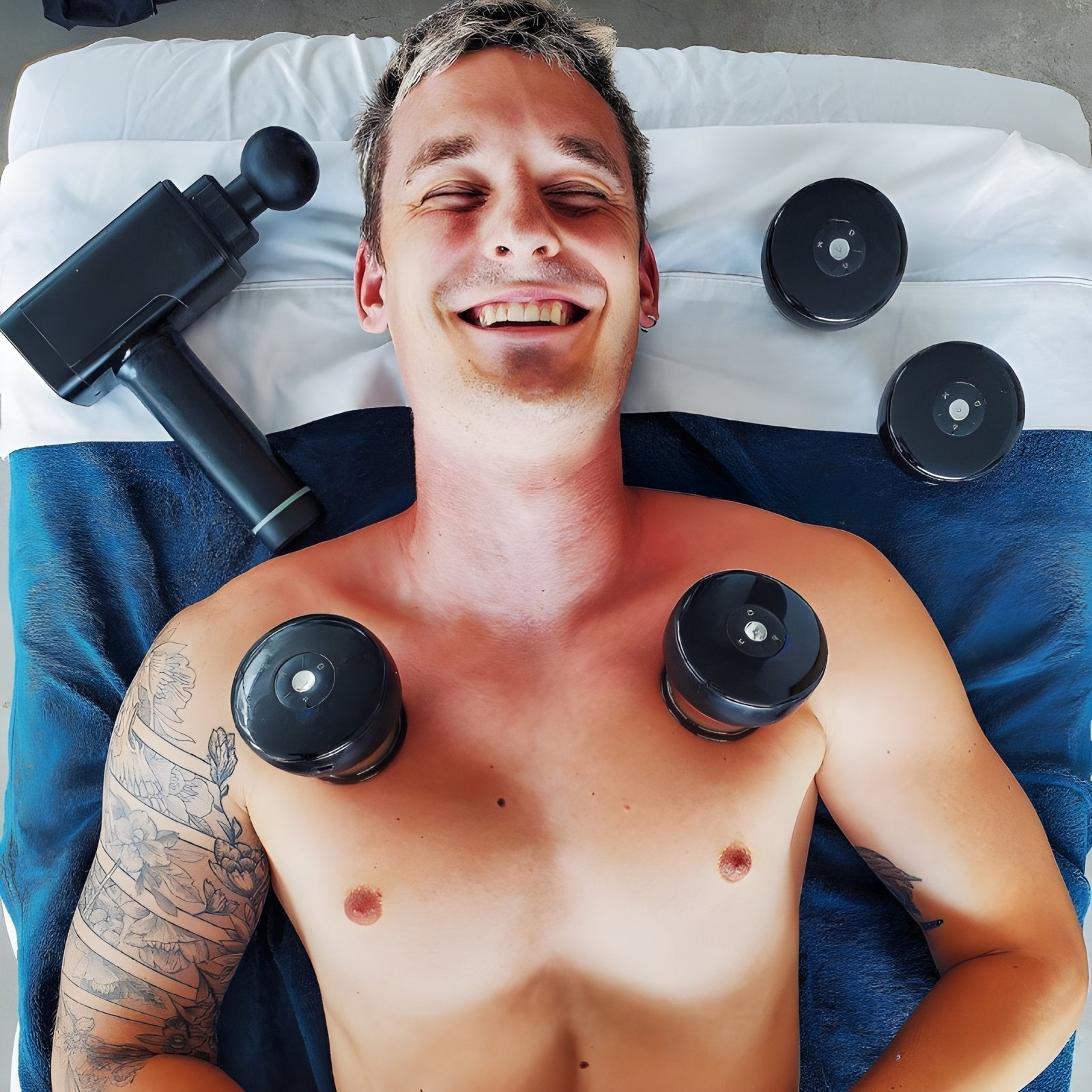 Lächelnder Mann bei Massage mit Massagepistole und Schröpfgläsern auf Brust, Tattoo am Arm.