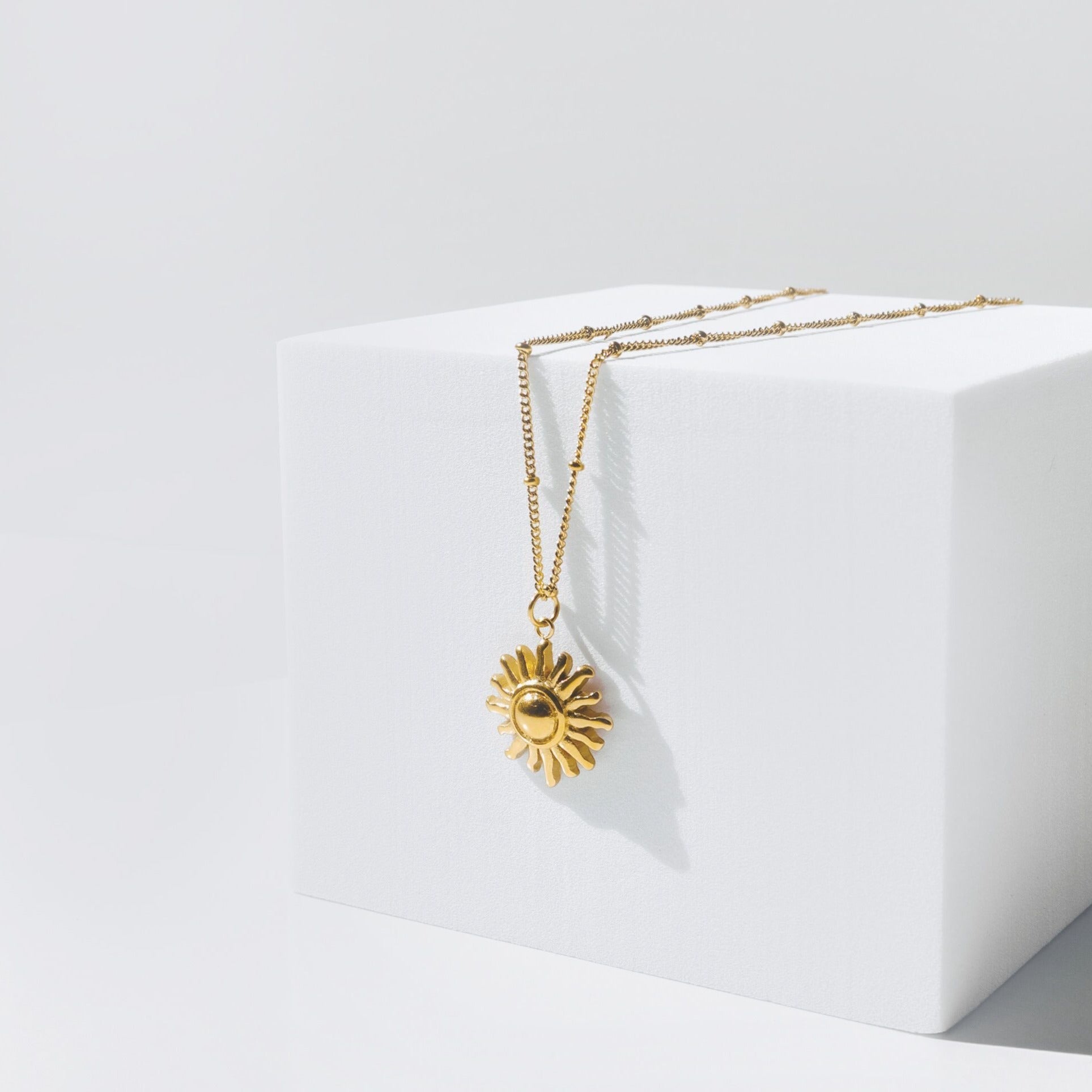 Goldene Sonnen-Halskette auf weißem Würfel, elegantes Schmuckdesign, modisches Accessoire.