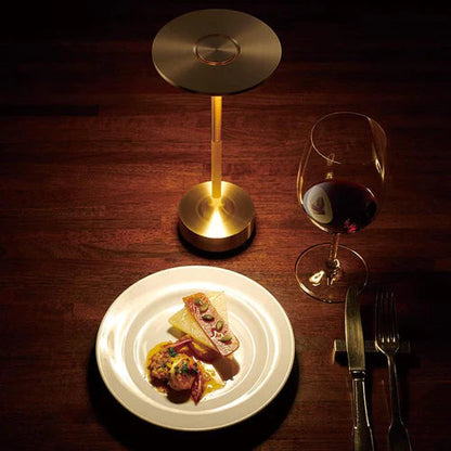 Goldene Tischlampe aus Metall, modernes Design, neben Teller mit Gourmet-Essen und Weinglas.
