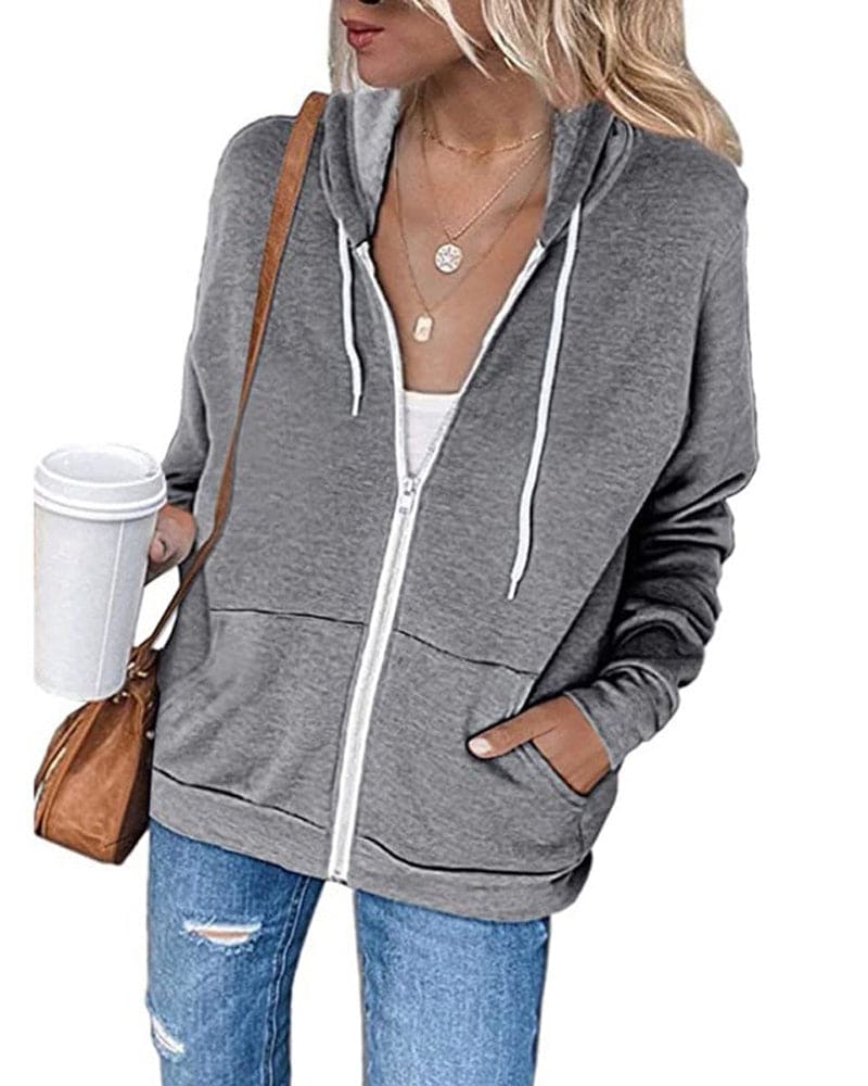 Frau in grauem Kapuzenpullover mit Reißverschluss, Kaffeebecher, Jeans, braune Tasche.