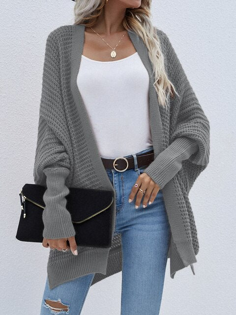 Frau in grauem Strickcardigan, weißem Top, blauer Jeans, hält schwarze Clutch.
