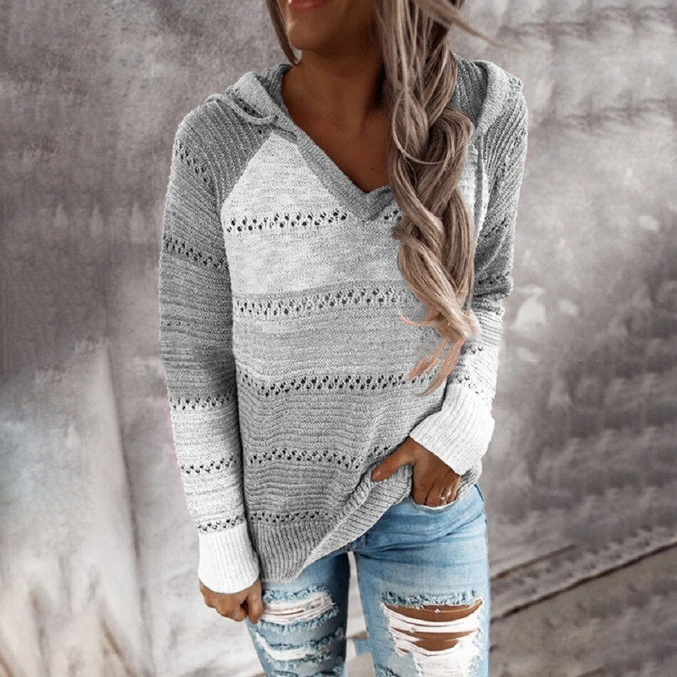 Frau in grauem Strickpullover mit Kapuze und zerrissenen Jeans vor Betonwand.