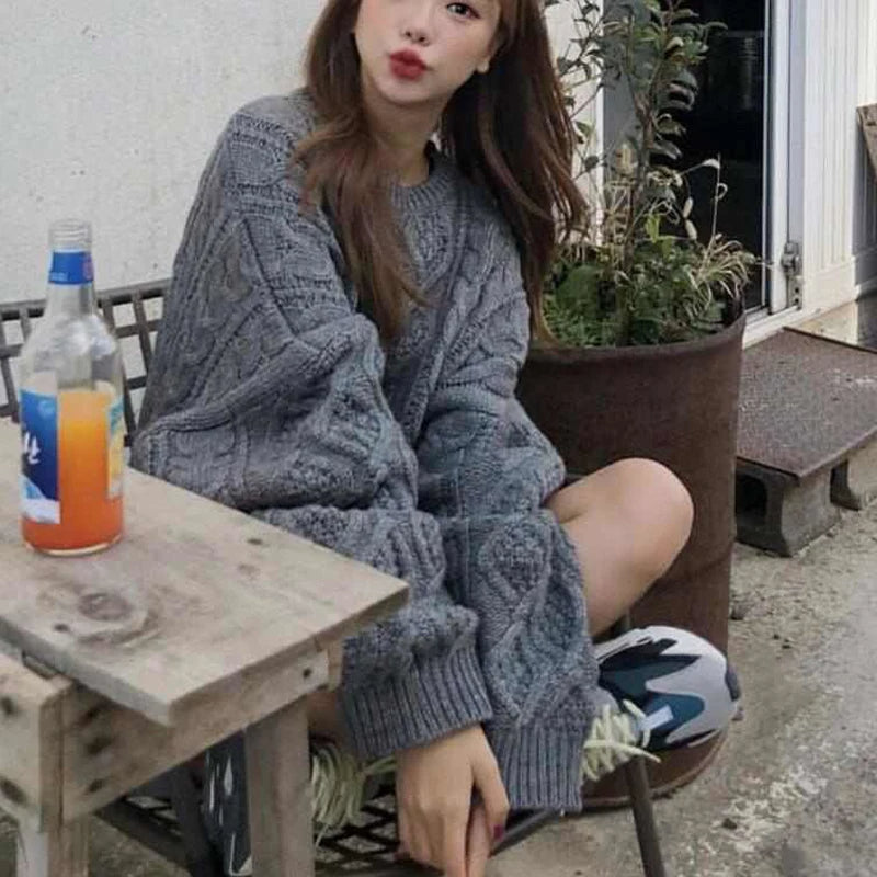 Frau in grauem Strickpullover sitzt draußen, neben Tisch mit Getränkeflasche, urbaner Hintergrund.