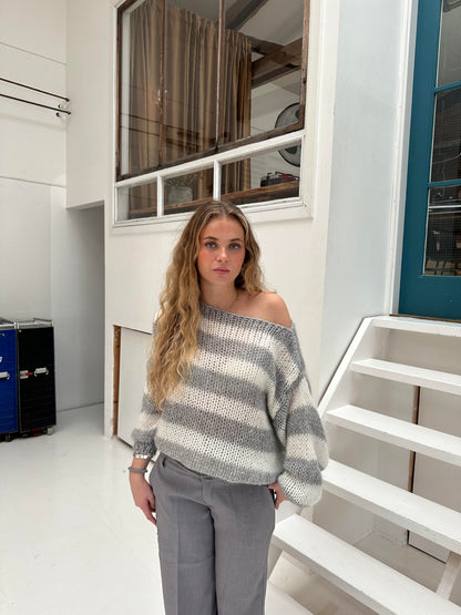 Frau in gestreiftem Pullover und grauer Hose steht vor weißer Treppe in modernem Innenraum.
