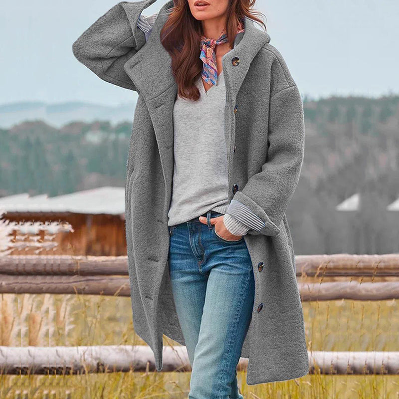 Frau in grauem Wollmantel, Jeans und Schal vor ländlicher Kulisse im Herbst.