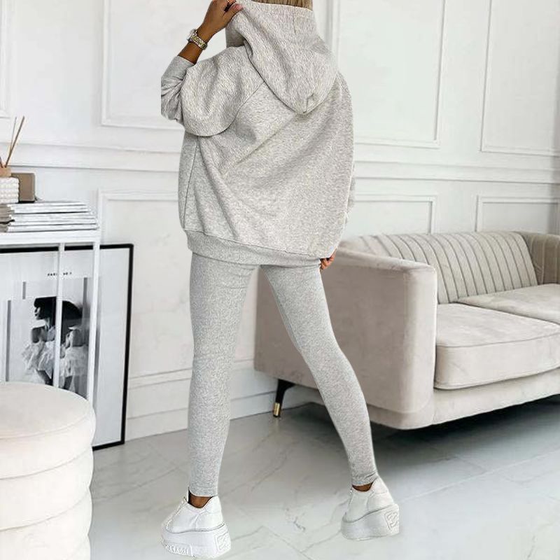 Frau in grauem Hoodie und Leggings, gemütliches Wohnzimmer, moderne Einrichtung, Mode-Outfit.