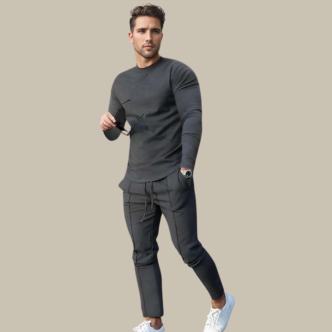 Mann in grauem Freizeitanzug mit Sonnenbrille und weißen Sneakern vor beigem Hintergrund.