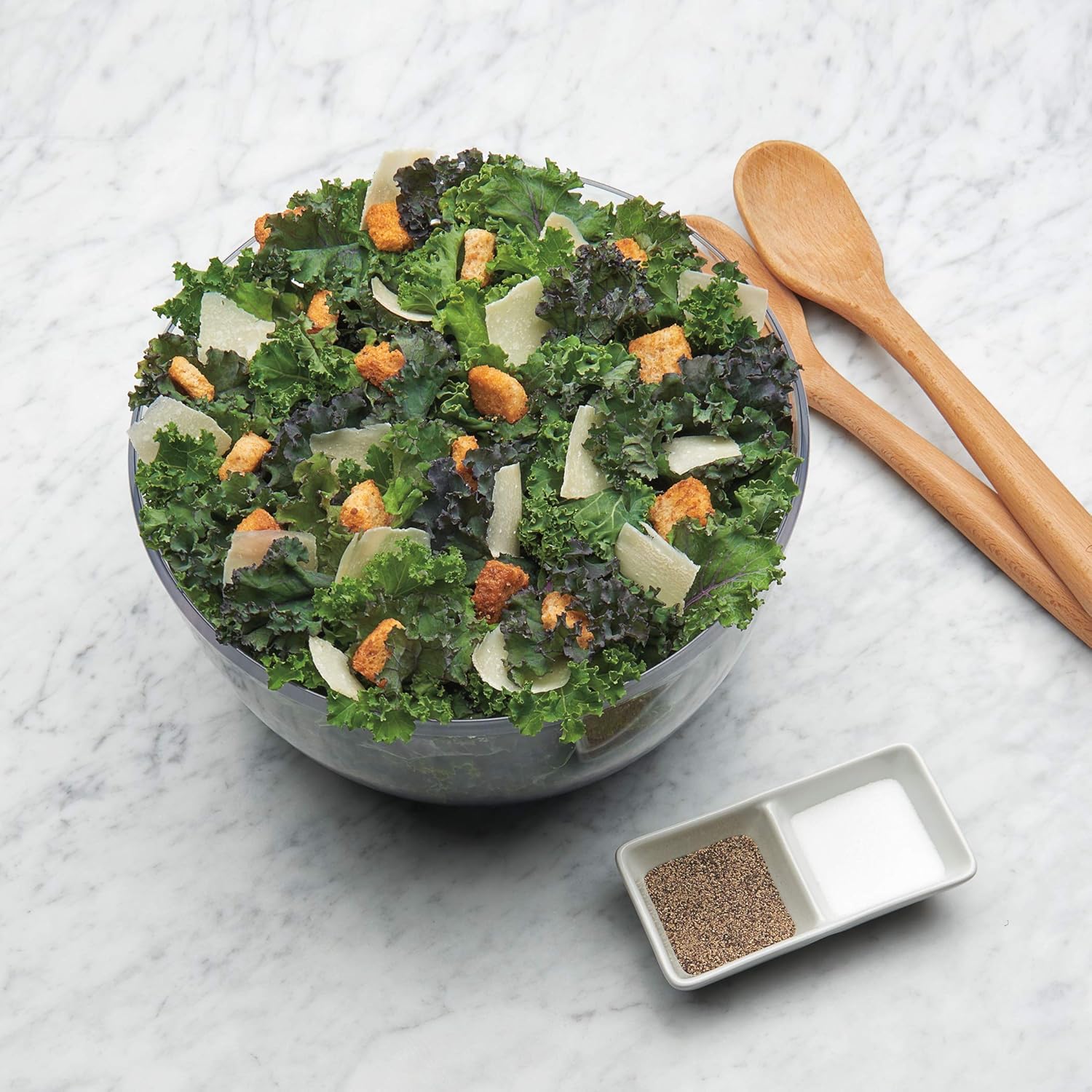 Grüner Salat mit Croutons und Parmesan in Glasschüssel auf Marmortisch, Holzlöffel daneben.