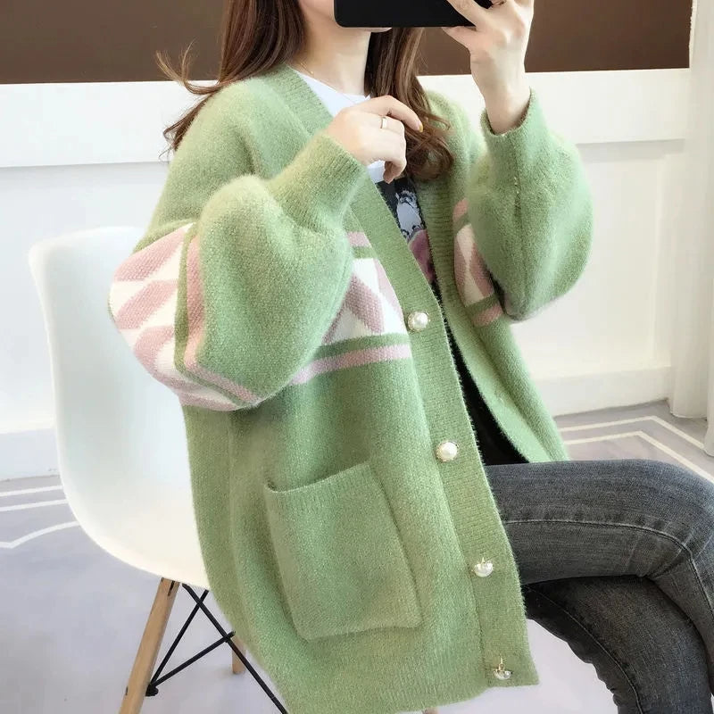 Frau trägt grüne Strickjacke mit rosa Muster, Knöpfen und Taschen, sitzt auf weißem Stuhl.