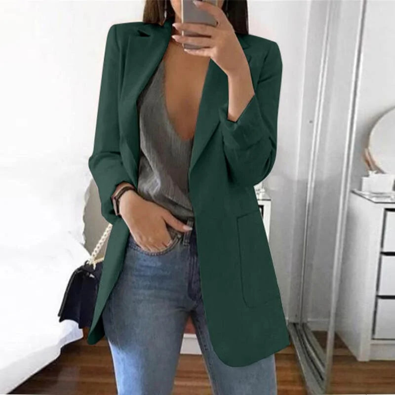 Frau in grünem Blazer, grauem Top, blauer Jeans, Spiegel-Selfie, modernes Schlafzimmer.