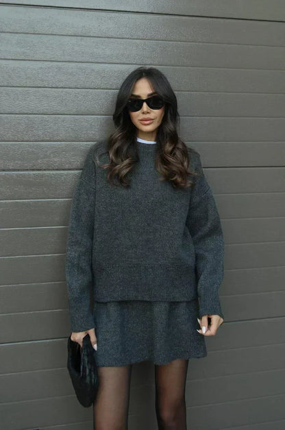 Model in anthrazitgrauem Strick-Set, Pullover mit breitem Saum und kurzer Rock, dazu Strumpfhose und schwarze Clutch. 11