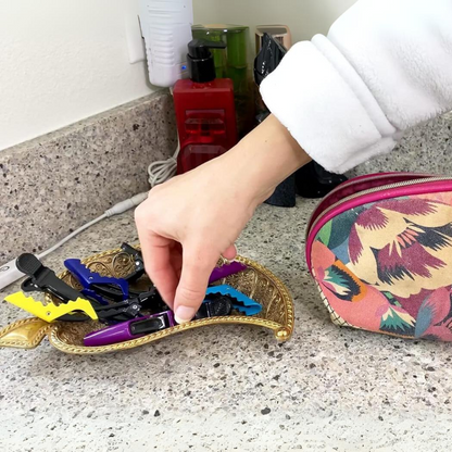 Hand greift nach bunten Haarklammern auf goldener Schale, bunte Kosmetiktasche daneben, Badezimmer.