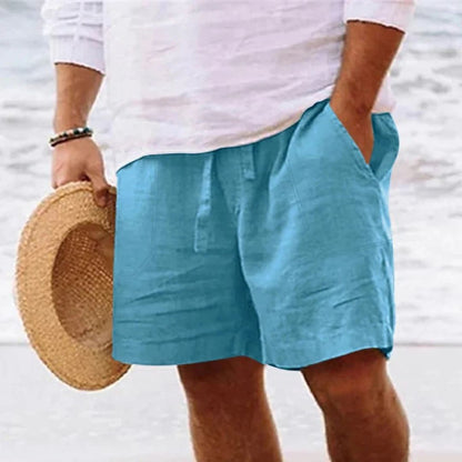Mann in blauer Leinen-Shorts und weißem Hemd am Strand, hält Strohhut.