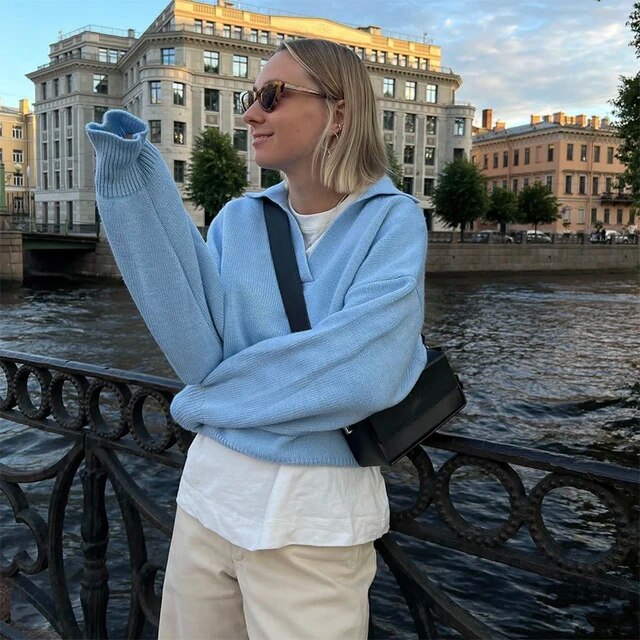 Frau in blauem Pullover und Sonnenbrille steht an Flussufer, Stadtgebäude im Hintergrund.