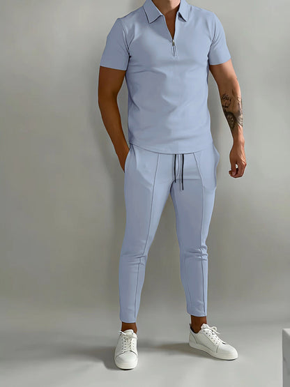 Mann in hellblauem Trainingsanzug und weißen Sneakers vor grauem Hintergrund.