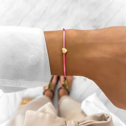 Dünnes pinkes Armband mit Herzanhänger, getragen am Handgelenk einer Person in weißer Bluse.