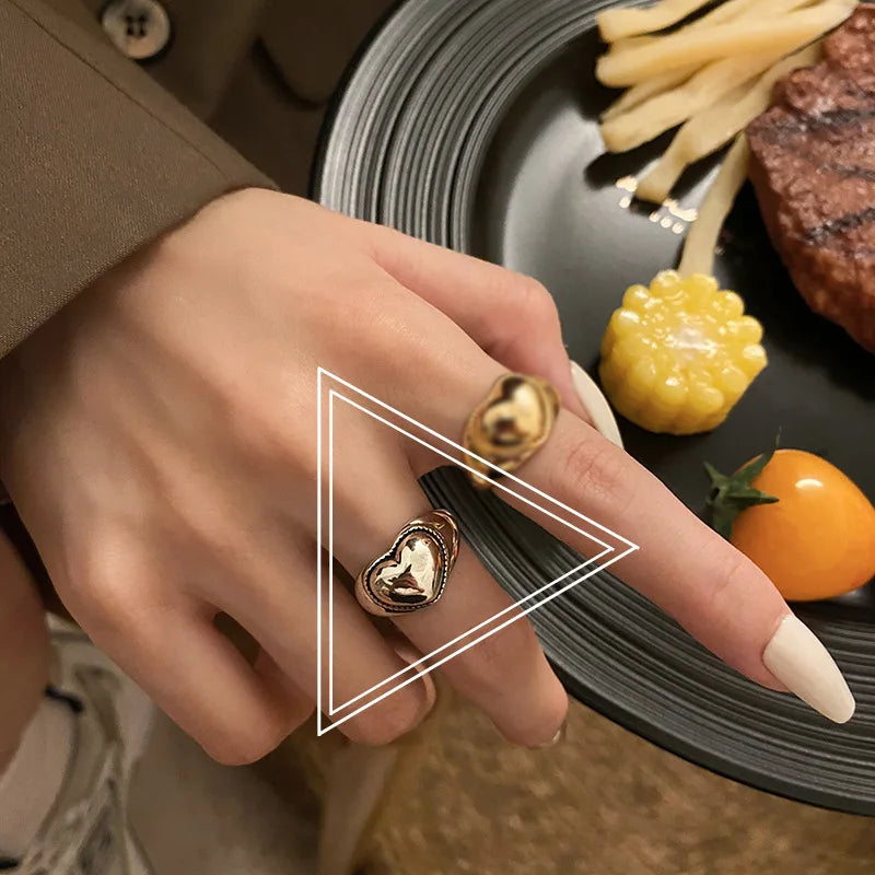 Damenhand mit goldfarbenem Herzring, Teller mit Pommes, Mais und Tomate im Hintergrund.