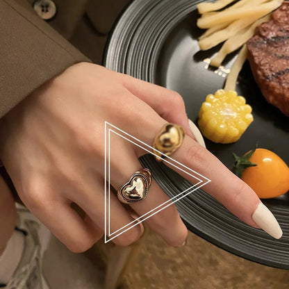Damenhand mit goldfarbenem Herzring, Teller mit Pommes, Mais und Tomate im Hintergrund.