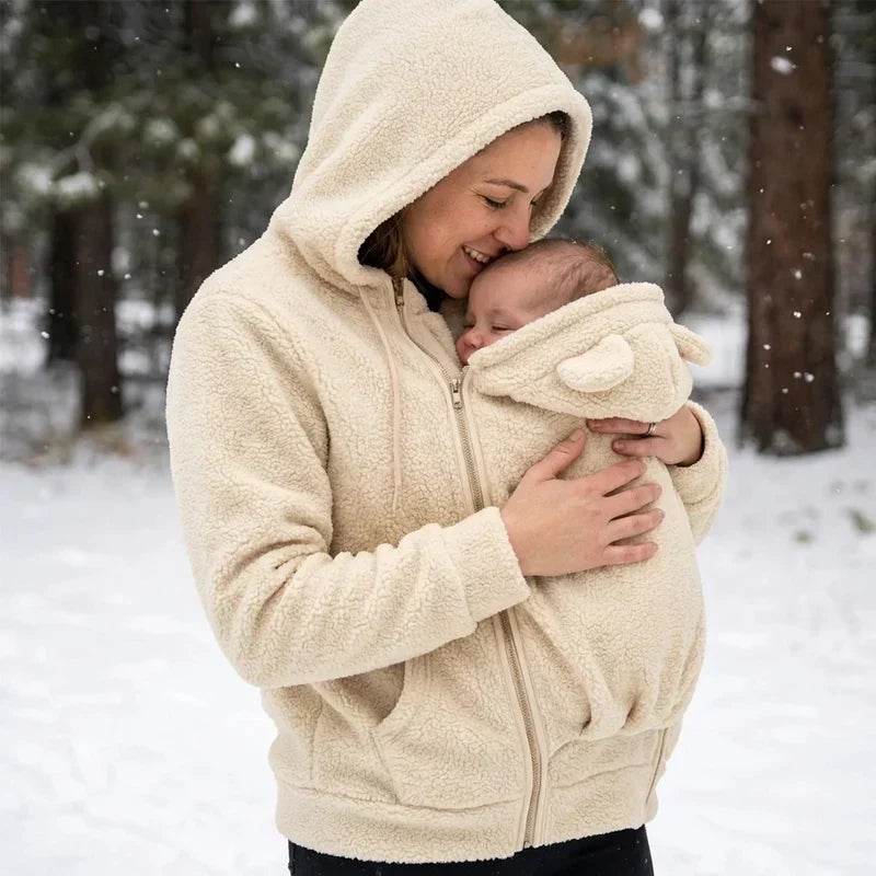 Frau im beigen Sherpa-Hoodie mit aufgesetzter Kapuze küsst ihr Baby im Trageeinsatz, verschneiter Wald im Hintergrund. 8