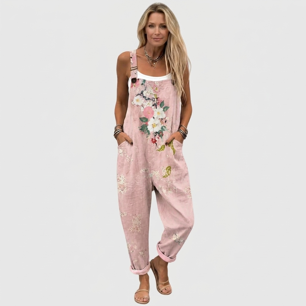 Frontansicht: Model mit langen blonden Haaren trägt die Latzhose mit Blumenmotiv, umgekrempelten Beinen und Sandalen, Hände in den Taschen. 2