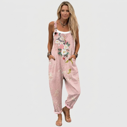 Frontansicht: Model mit langen blonden Haaren trägt die Latzhose mit Blumenmotiv, umgekrempelten Beinen und Sandalen, Hände in den Taschen. 2