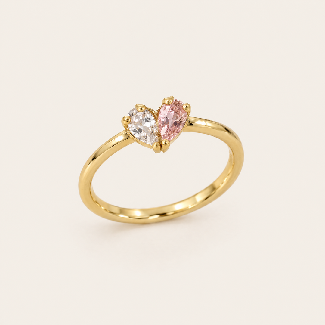 Goldfarbener Ring in Schrägansicht, dessen zwei tropfenförmige Steine, ein klarer und ein rosafarbener, gemeinsam ein Herz bilden. 1
