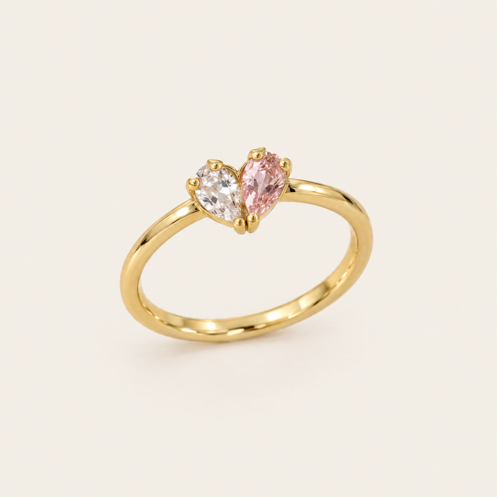 Goldfarbener Ring in Schrägansicht, dessen zwei tropfenförmige Steine, ein klarer und ein rosafarbener, gemeinsam ein Herz bilden. 1
