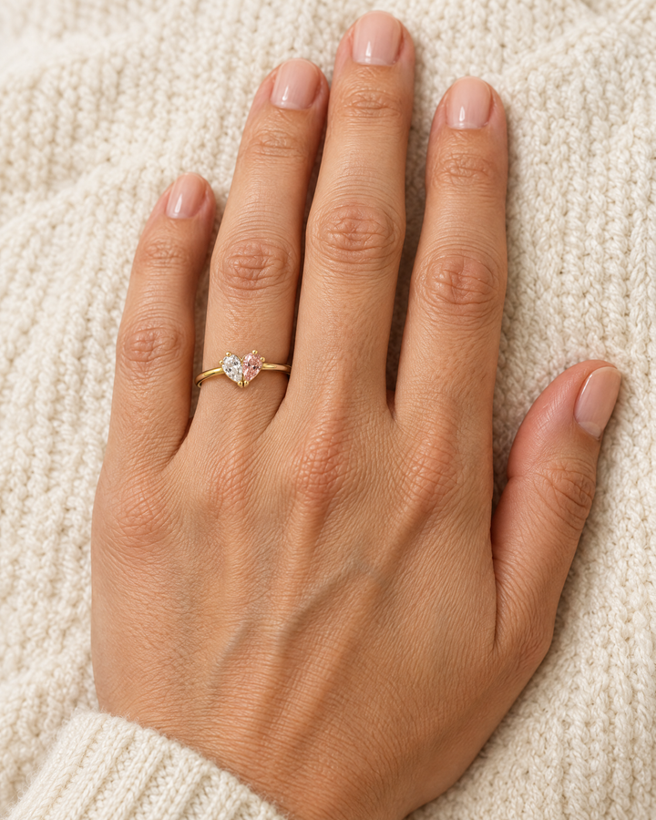 Der Ring mit klarem und rosafarbenem Stein am Finger einer Hand, die auf einem cremefarbenen Strickstoff liegt. 2