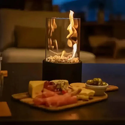 Tischkamin mit Glaszylinder, brennend, auf Holztisch neben Käseplatte und Oliven, gemütliche Atmosphäre.