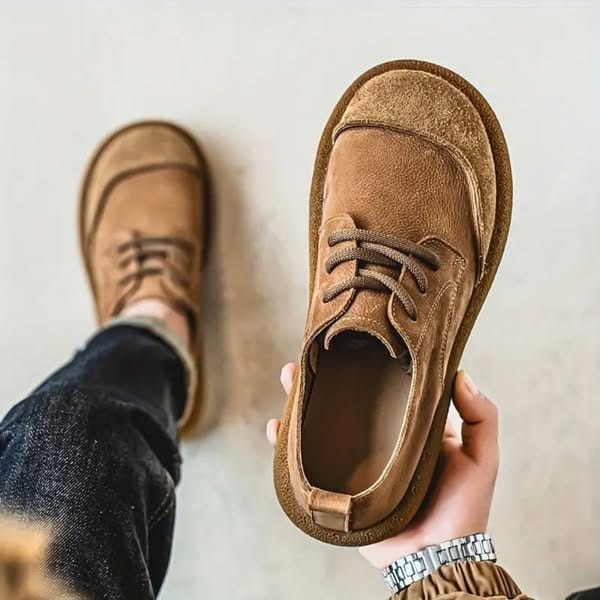 Hellbrauner Schnürschuh in Wildleder-Optik wird von einer Hand mit Armbanduhr gehalten, im Hintergrund trägt jemand das zweite Paar in Jeans. 1