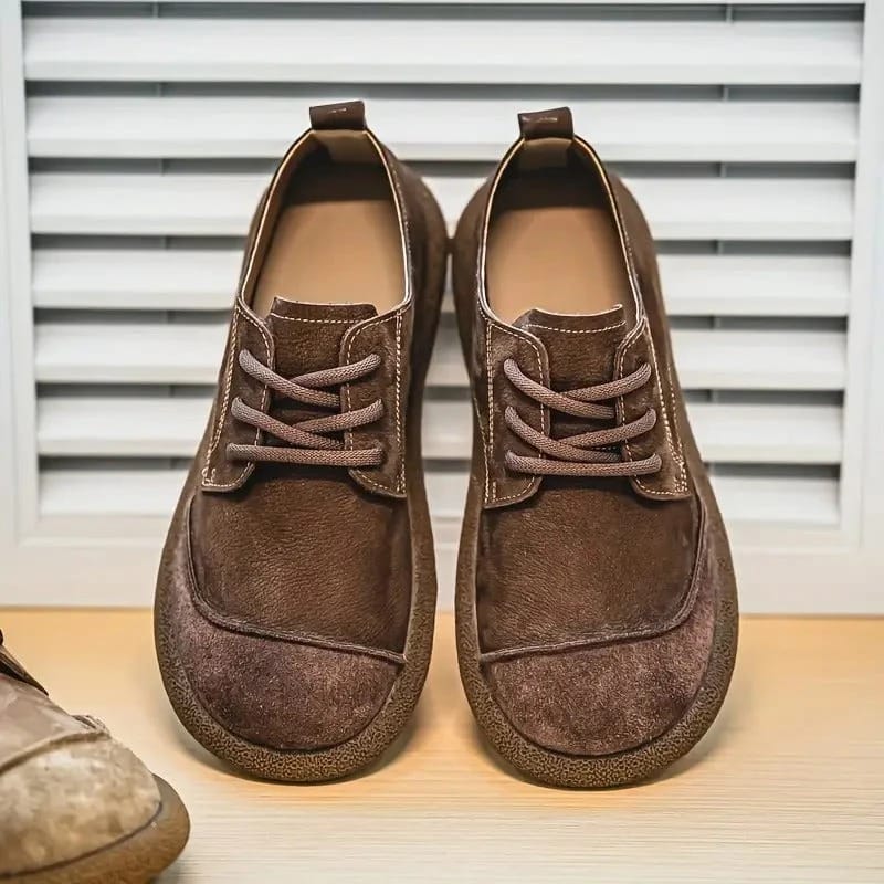 Paar dunkelbraune Schnürschuhe von vorn auf einem Holzbrett vor einer Lamellenwand, links ragt ein hellerer Schuh ins Bild. 6