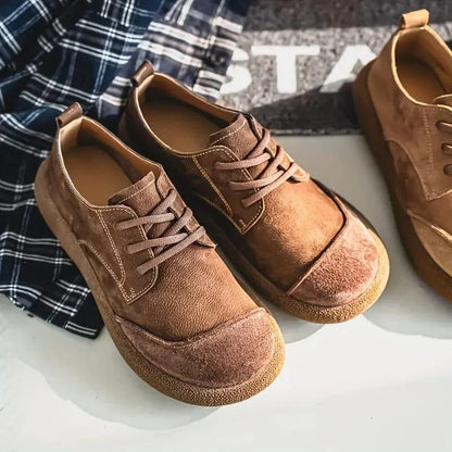 Zwei braune Schnürschuhe mit heller Naht und Kappe schräg auf weißem Untergrund neben einem karierten Stoff, ein dritter Schuh am Rand. 11
