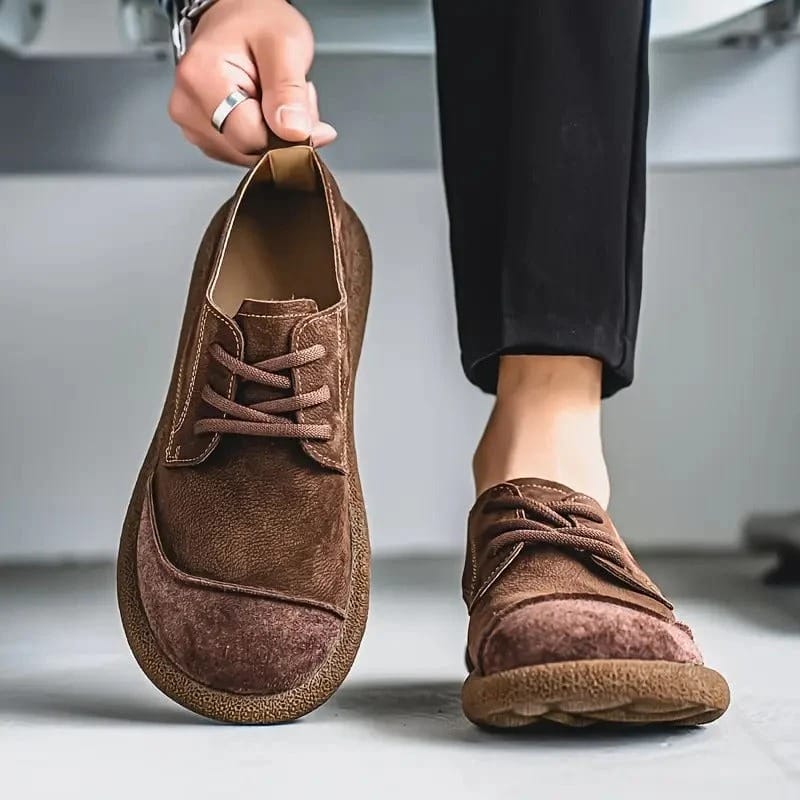 Dunkelbrauner Schnürschuh wird an der Fersenlasche hochgehalten, der zweite Schuh am Fuß neben schwarzer Hose, breite Gummisohle sichtbar. 8