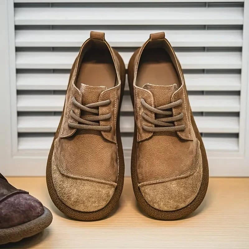 Frontansicht eines hellbraunen Schuhpaars mit Schnürung und aufgerauter Kappe auf Holzfläche vor weißer Lamellenwand. 9