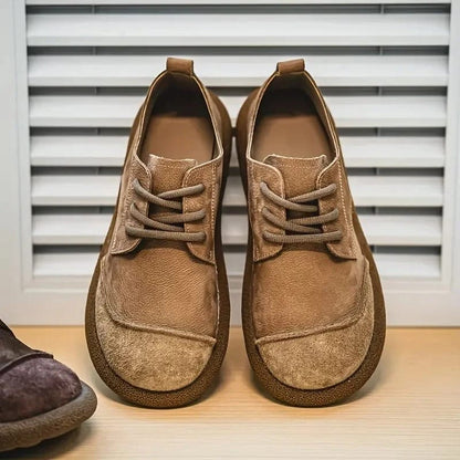 Frontansicht eines hellbraunen Schuhpaars mit Schnürung und aufgerauter Kappe auf Holzfläche vor weißer Lamellenwand. 9