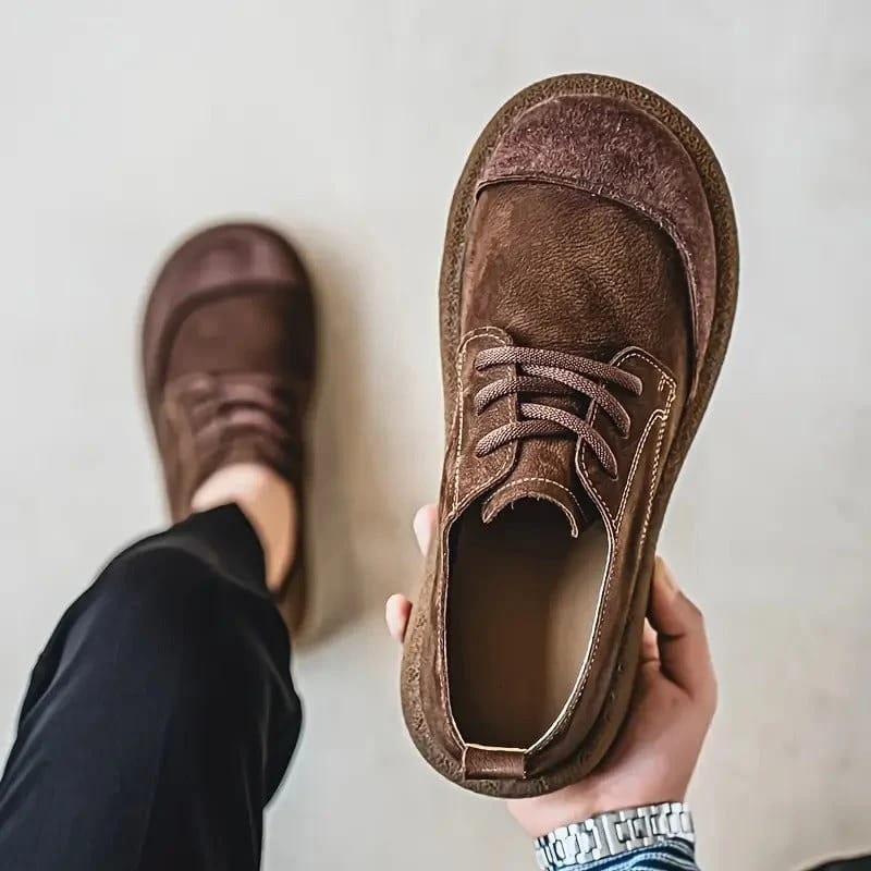 Dunkelbrauner Schnürschuh mit Kontrastnähten wird von oben in der Hand gehalten, daneben ein getragener Schuh zur schwarzen Hose. 2