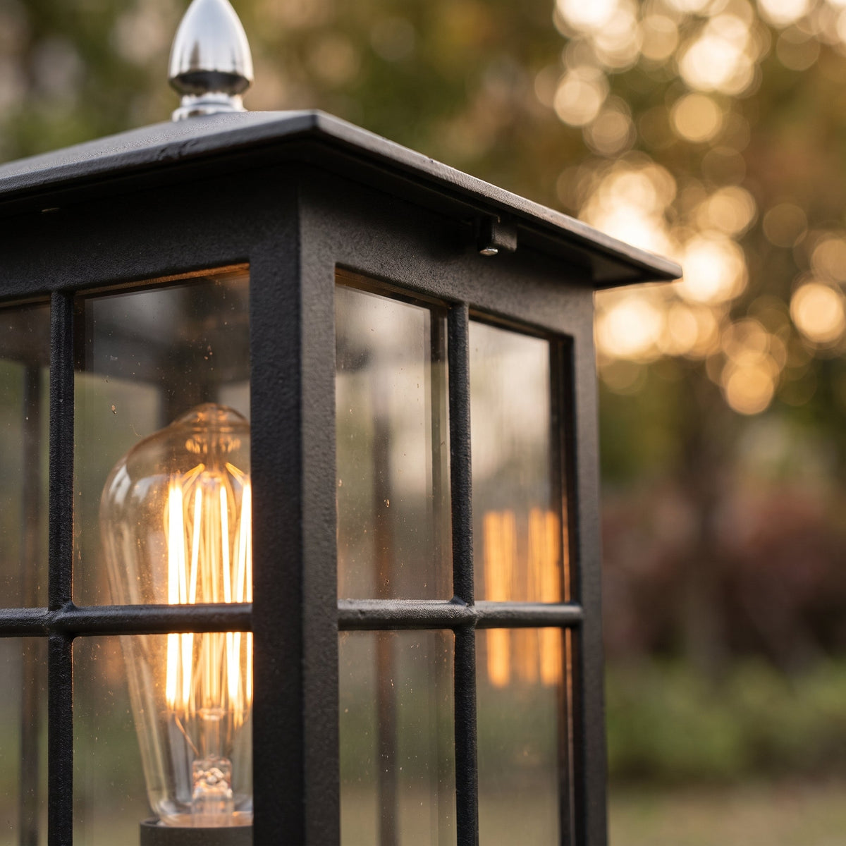 Nahaufnahme des schwarzen Metallrahmens mit strukturierter Oberfläche, Glasscheiben, glühender Lampe und silberfarbener Dachspitze vor sonnigem Gartenhintergrun 5