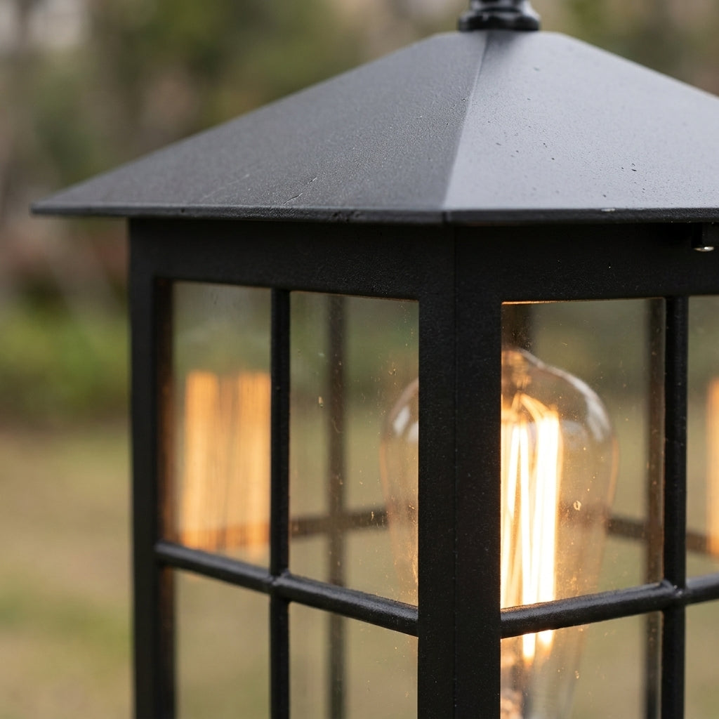 Detailaufnahme des schwarzen Pyramidendachs und der Sprossenfenster der Leuchte, im Inneren eine warm leuchtende Glühfadenlampe, Garten unscharf im Hintergrund. 6