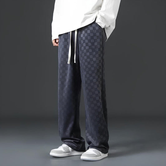 Mann in karierten, dunkelgrauen Jogginghosen und weißen Sneakers vor grauem Hintergrund.